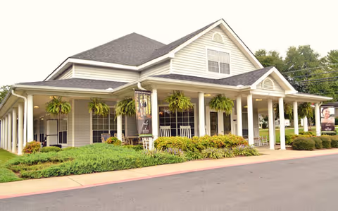 Exterior view of a single-story building with a large covered porch supported by white columns. The building has beige siding, a dark gray roof, and several hanging green plants along the porch. There are shrubs and greenery in front of the porch and a paved driveway or road in the foreground.