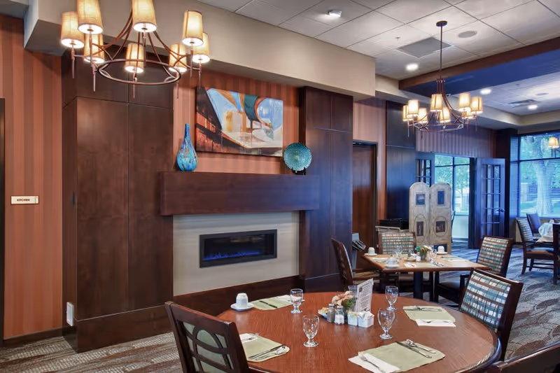 Dining room with round tables set for a meal, a wall-mounted fireplace, chandeliers, and large windows.