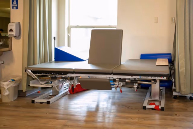 A medical examination or therapy table with adjustable sections in a room with wood flooring, a window with curtains, a wall-mounted hand sanitizer dispenser, and a trash bin.