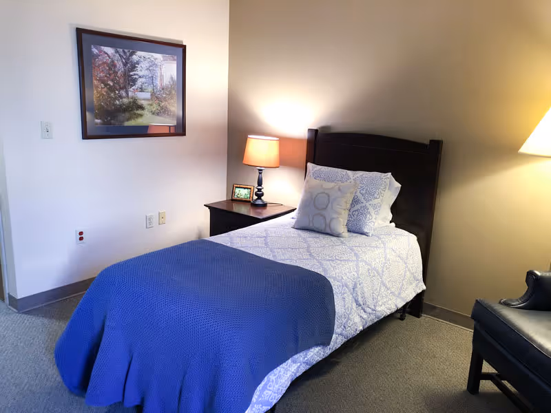A neatly made single bed with patterned white and blue bedding and a blue blanket at the foot. Next to the bed is a dark wooden nightstand with a lit table lamp and a small framed picture. On the wall above the nightstand is a framed painting of a garden scene. To the right of the bed is a dark upholstered chair. The room has beige walls and carpeted floor.