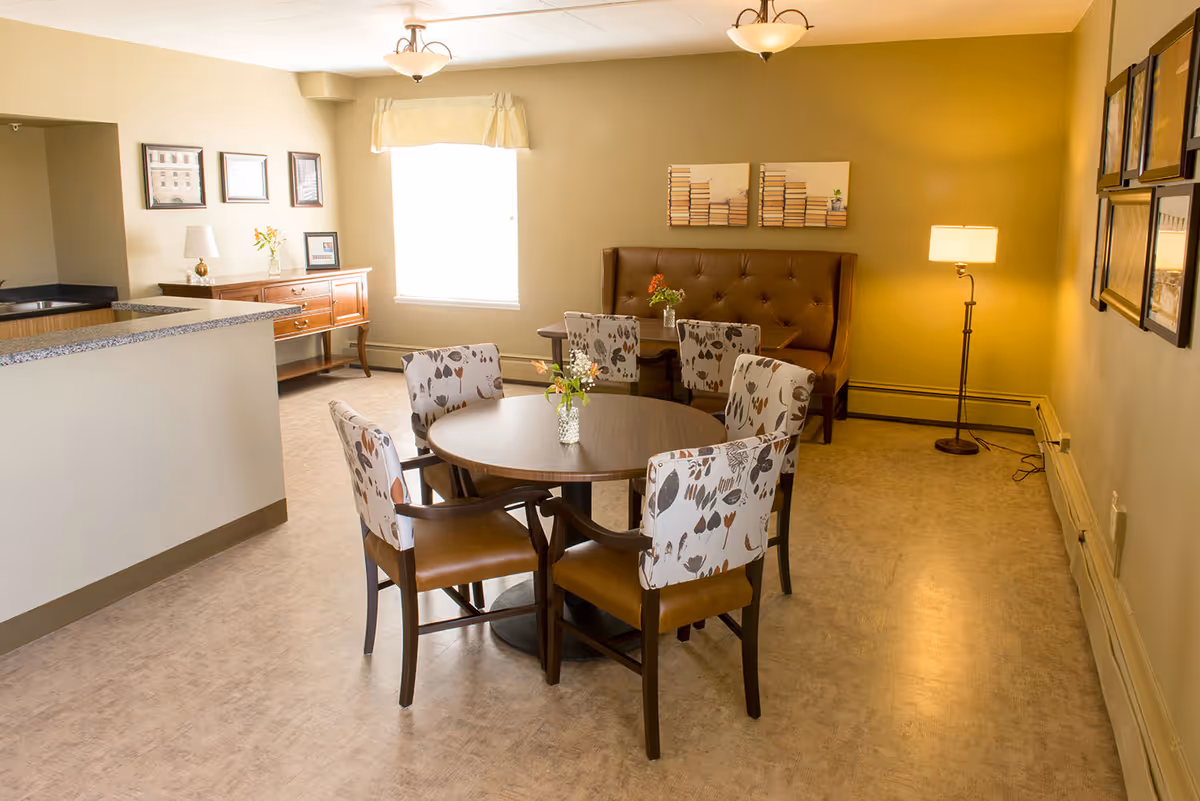 A cozy dining area in a senior living facility with a round wooden table surrounded by six chairs, four with floral patterned upholstery and two with brown leather seats. There is a cushioned bench along the back wall with a small table in front of it. The room is warmly lit with ceiling lights and a floor lamp, and decorated with framed pictures and a sideboard with a lamp and flowers near a window with a valance.