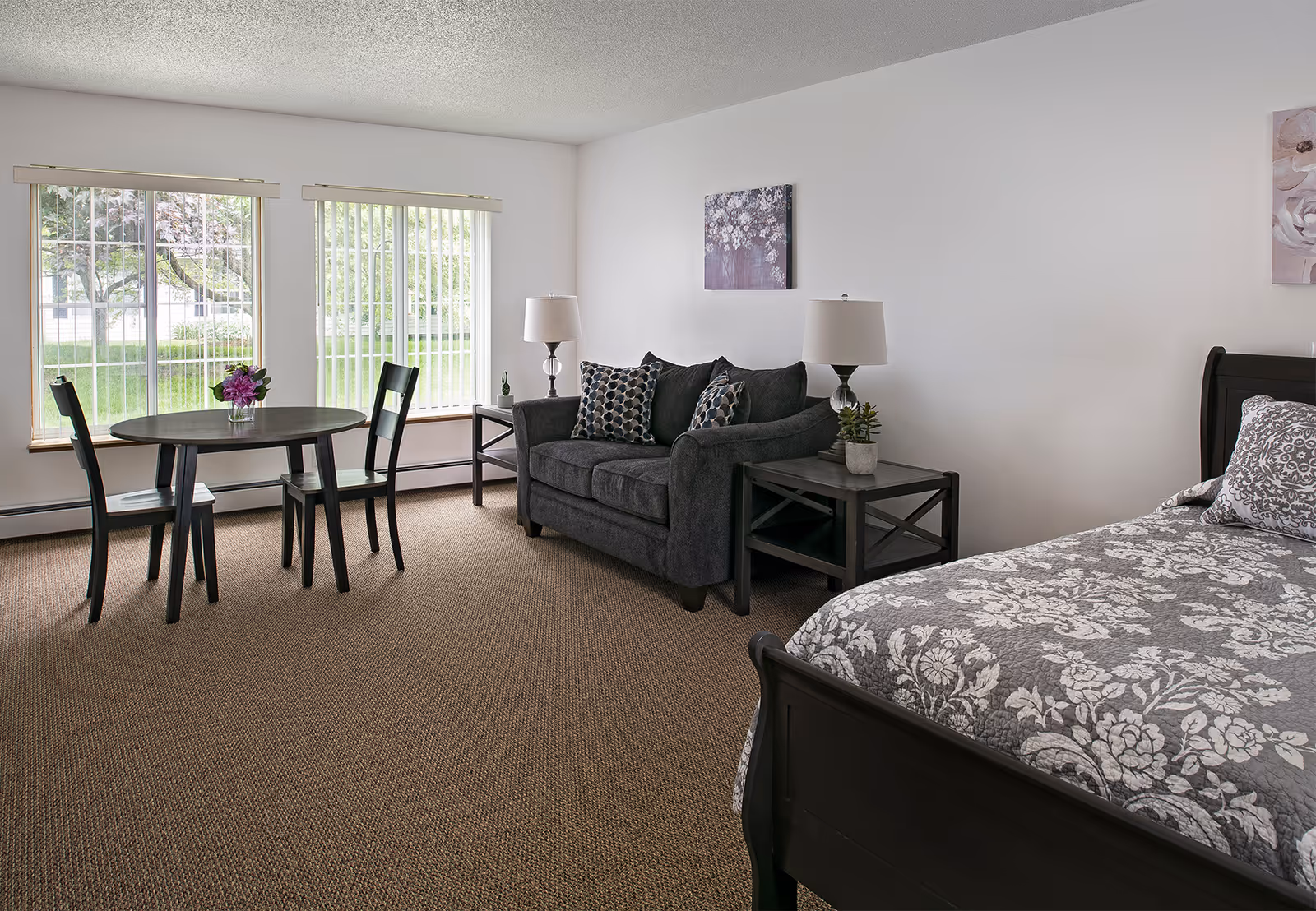 A bright and cozy room featuring a small round dining table with two chairs near two large windows with vertical blinds. A dark gray loveseat with patterned pillows sits against a white wall, flanked by two side tables each with a lamp and small plants. Part of a bed with a floral patterned gray and white bedspread is visible in the foreground.