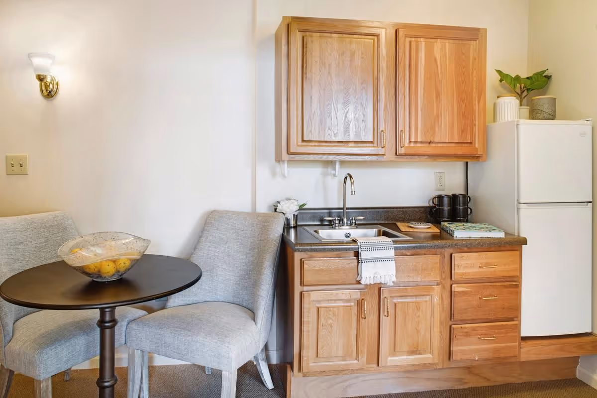 A small kitchen area with wooden cabinets, a sink with a towel hanging on the faucet, a countertop with a few items including a book and mugs, and a white refrigerator. Next to the kitchen is a small round table with two gray upholstered chairs and a decorative bowl with fruit on the table. A wall sconce light is mounted on the wall above the chairs.