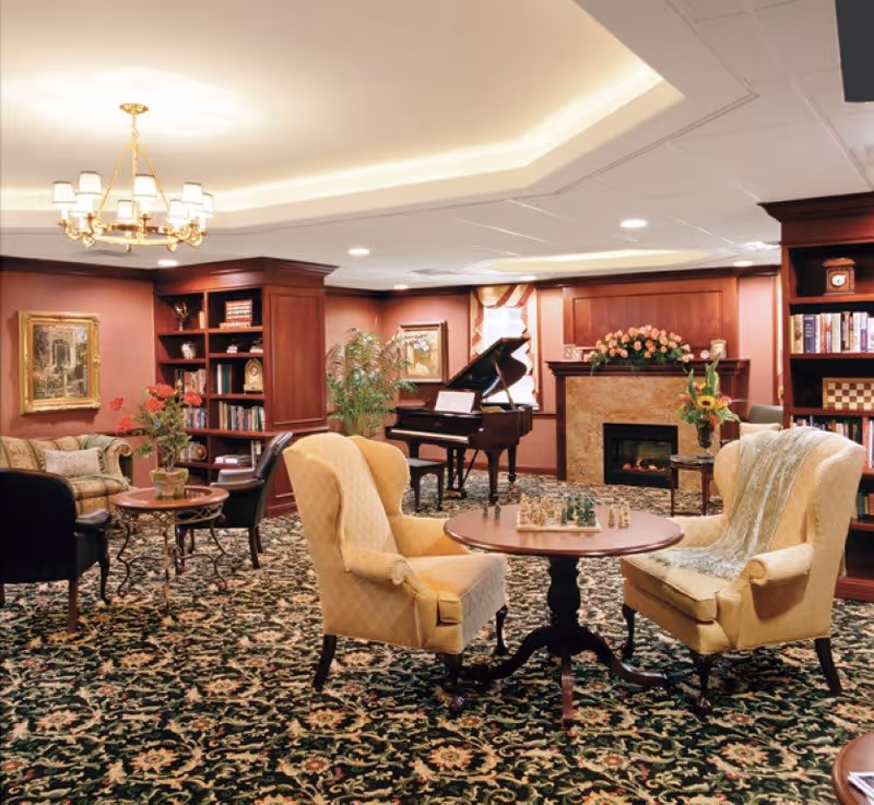 A cozy living room with patterned carpet, two yellow armchairs facing a round wooden table with a chess set, a grand piano near a window, a fireplace with floral decoration, bookshelves filled with books and decorative items, and a chandelier hanging from the ceiling.