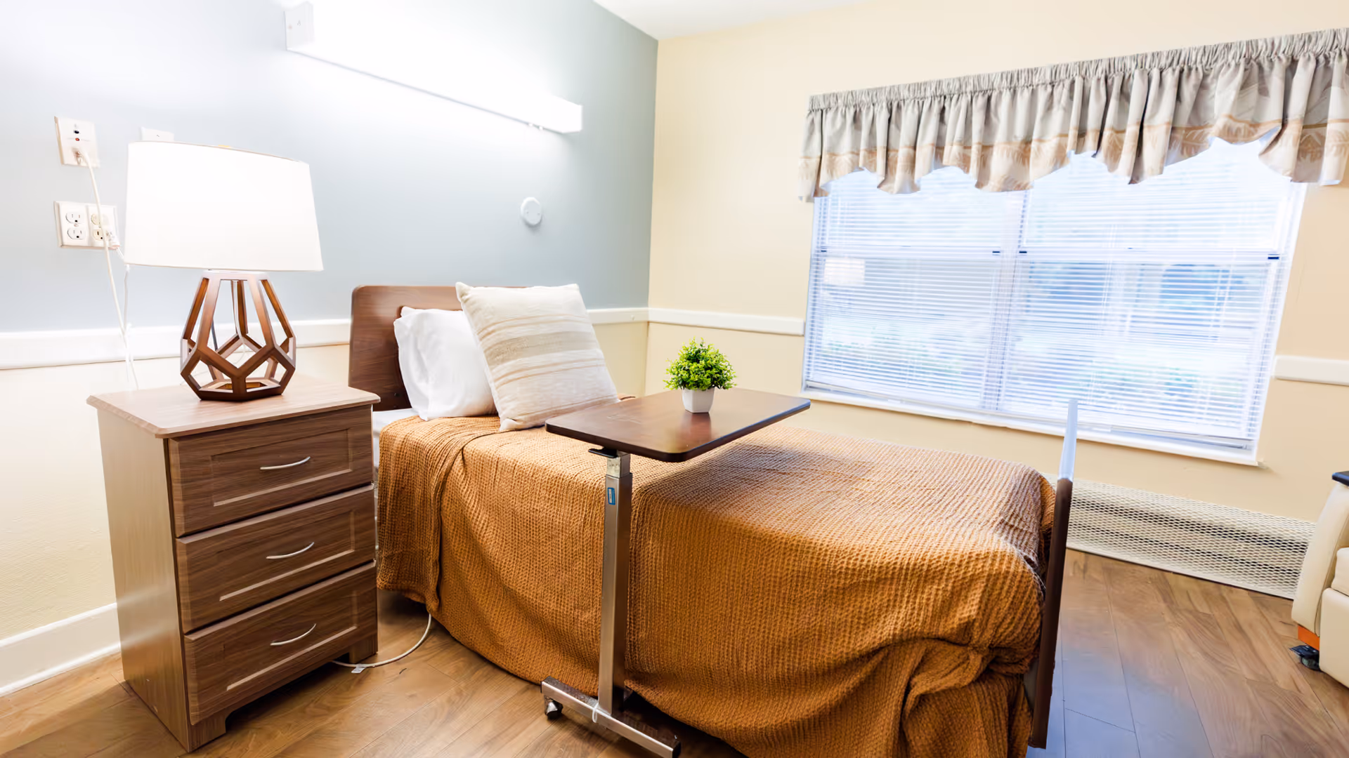 A neatly made single bed with a brown blanket and two pillows in a well-lit room. Next to the bed is a wooden nightstand with three drawers and a modern lamp on top. A small overbed table with a small green plant is positioned over the bed. The room has a large window with blinds and a valance, and the floor is wooden.