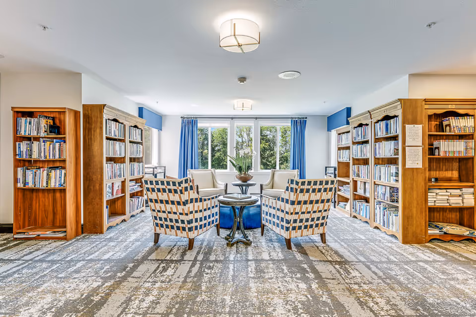 A bright and spacious library room with large windows letting in natural light. The room features wooden bookshelves filled with books along the walls, two patterned armchairs, two white armchairs, a round blue ottoman, and a small round table with a plant in the center. Blue curtains frame the windows, and modern ceiling lights illuminate the space.