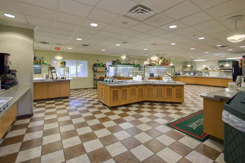 Spacious dining area with checkered tile floor, multiple food service counters with glass sneeze guards, and a coffee station on the left. The room has a drop ceiling with recessed lighting and large windows letting in natural light.