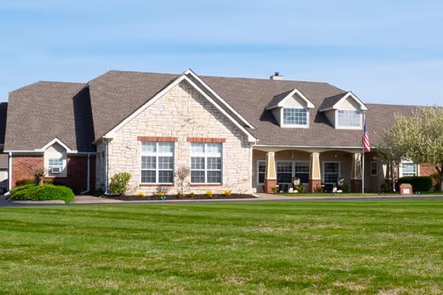 Front exterior view of a single-story senior living facility building with a stone and brick facade, multiple windows, a covered porch with columns, an American flag, and a well-maintained green lawn in front under a clear blue sky.