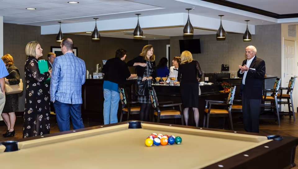 A group of people socializing in a lounge area with a pool table in the foreground. The room has pendant lights hanging from the ceiling, a bar counter with drinks, and several chairs around. The setting appears to be a common area in a senior living facility.