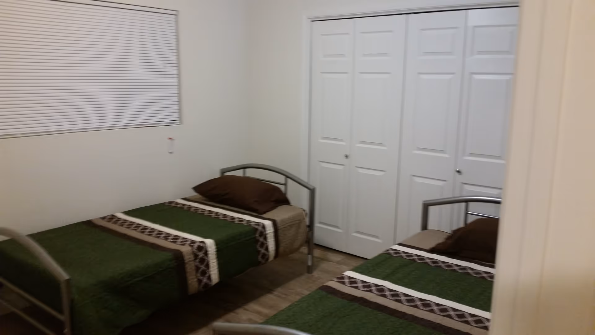 A small bedroom with two single beds, each with a green, brown, and beige patterned bedspread and a brown pillow. The room has white walls, a window with closed blinds, and a large white closet with double doors.