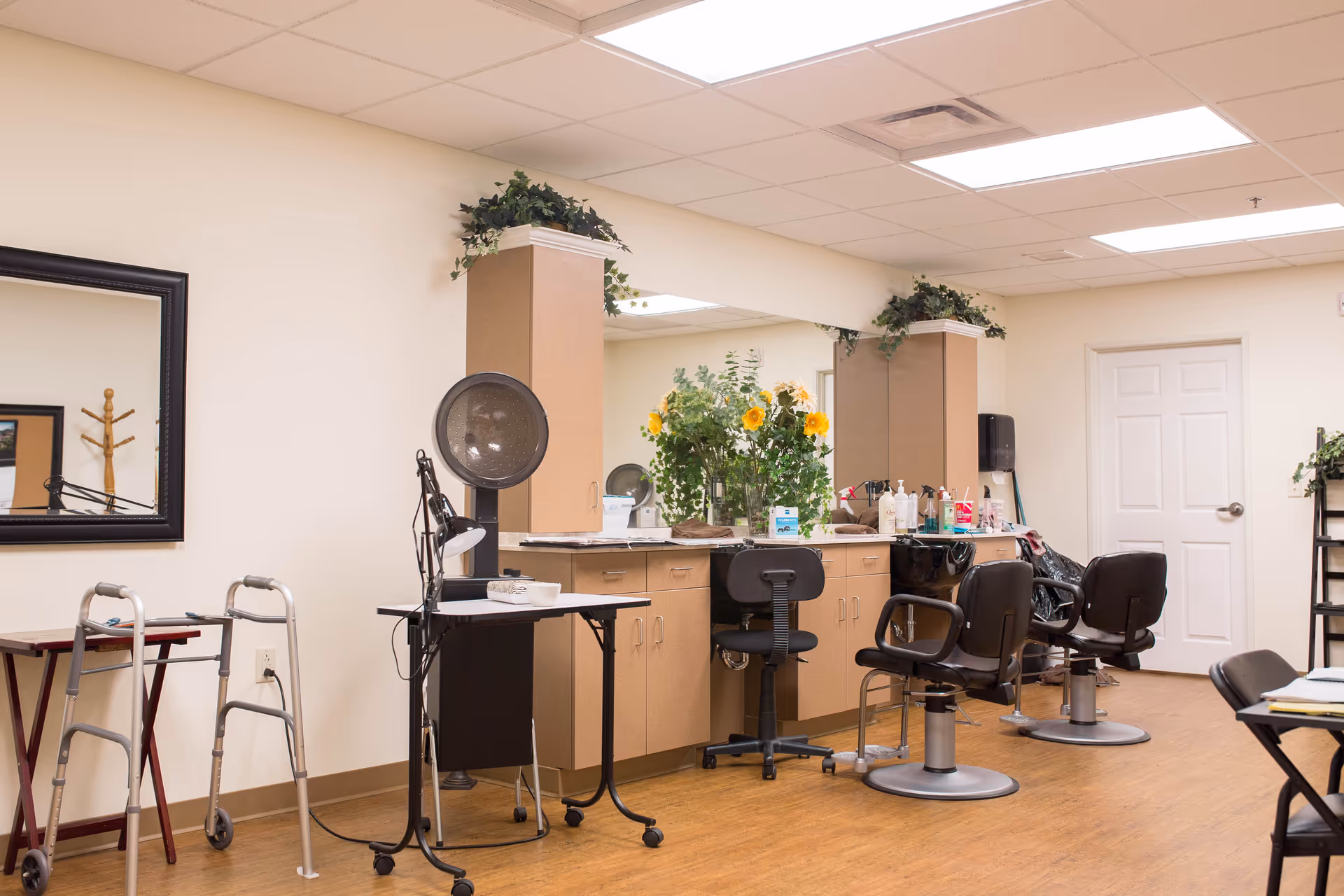Interior view of a salon area in a senior living facility with salon chairs, a hair dryer, a walker, a large mirror, cabinets with various hair and beauty products, and decorative plants including sunflowers.