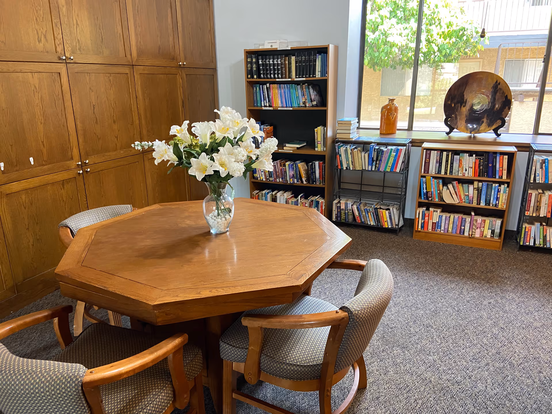 A cozy room with a wooden octagonal table surrounded by four cushioned chairs. On the table is a clear vase filled with white flowers. The room has multiple bookshelves filled with books, a large window letting in natural light, and wooden cabinets along one wall.
