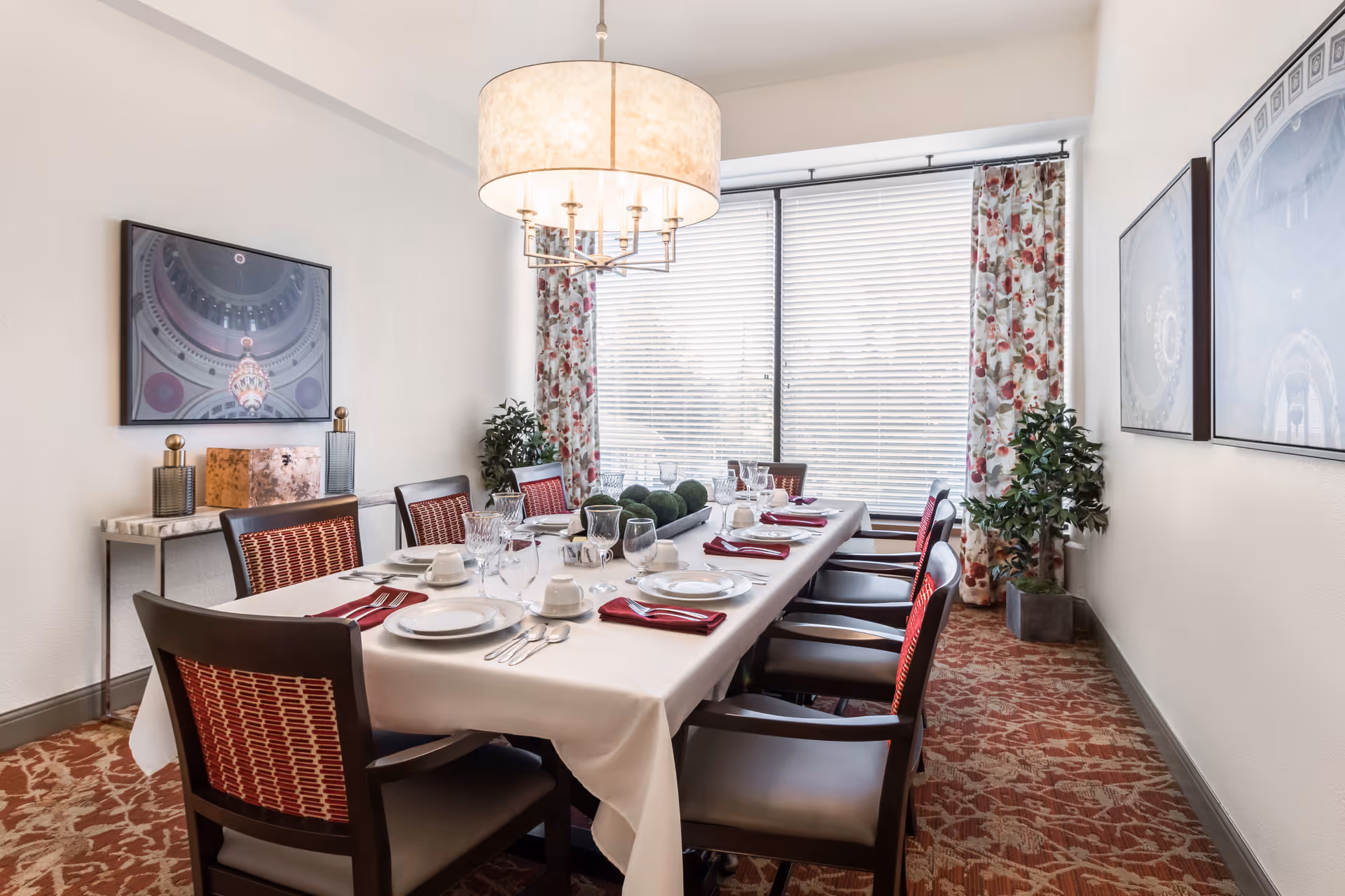 A formal dining room with a long table set for a meal, a chandelier overhead, chairs, and a large window with blinds and floral curtains.