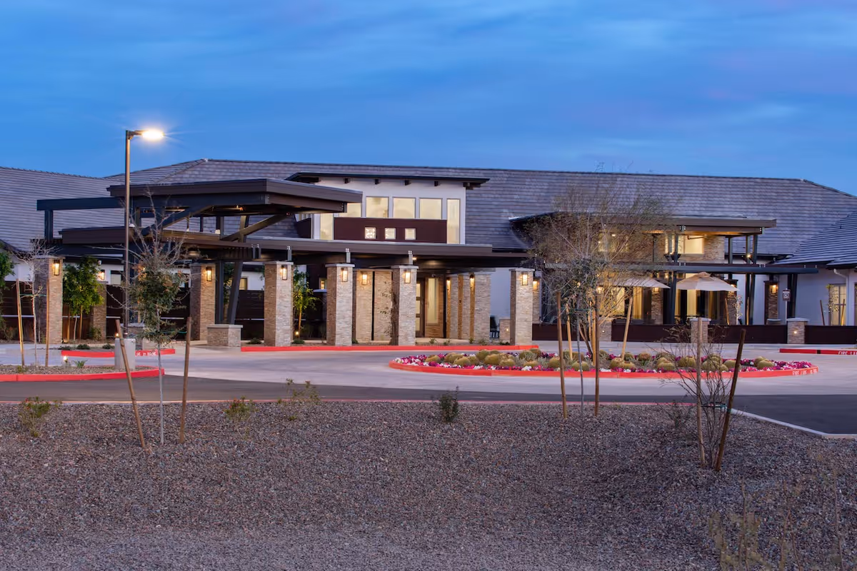 Exterior view of Spring Gardens Senior Living Peoria at dusk, showing the building entrance with stone pillars, a covered drop-off area, landscaped roundabout with flowers and cacti, and a lit streetlamp.
