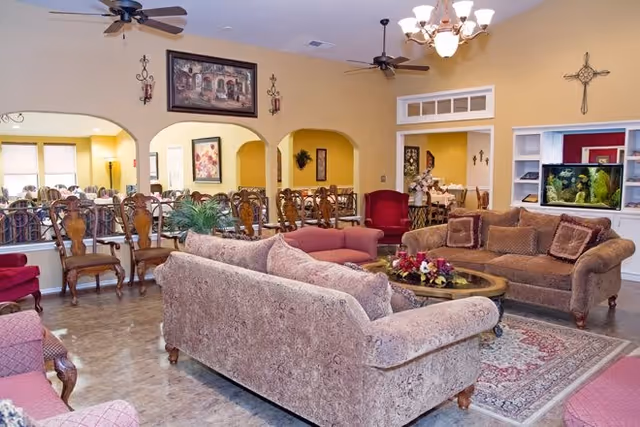 A spacious living room area in a senior living facility with multiple upholstered sofas and armchairs arranged around a central coffee table with a floral centerpiece. The room features a chandelier, ceiling fans, decorative wall art, and a built-in aquarium. In the background, there is a dining area with wooden chairs and tables visible through arched openings.