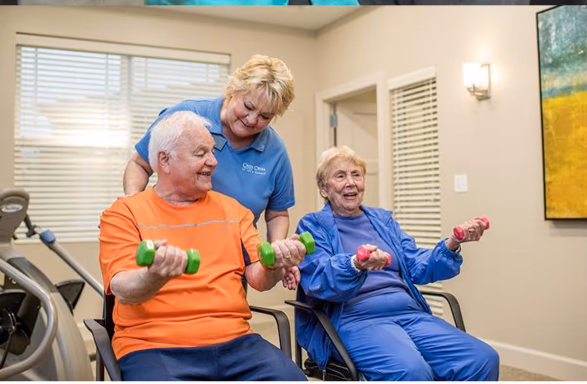 Two elderly individuals seated and holding small dumbbells while a caregiver in a blue shirt stands behind them, assisting and smiling. The setting appears to be a well-lit indoor room with exercise equipment and a large window with blinds.