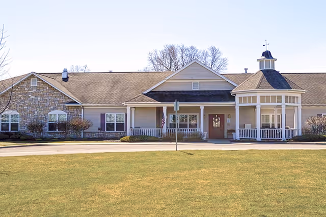 Exterior view of a single-story senior living facility building with a stone and siding facade, a covered entrance with white columns, and a small cupola on the roof. There is a well-maintained lawn in front and some leafless trees in the background under a clear sky.