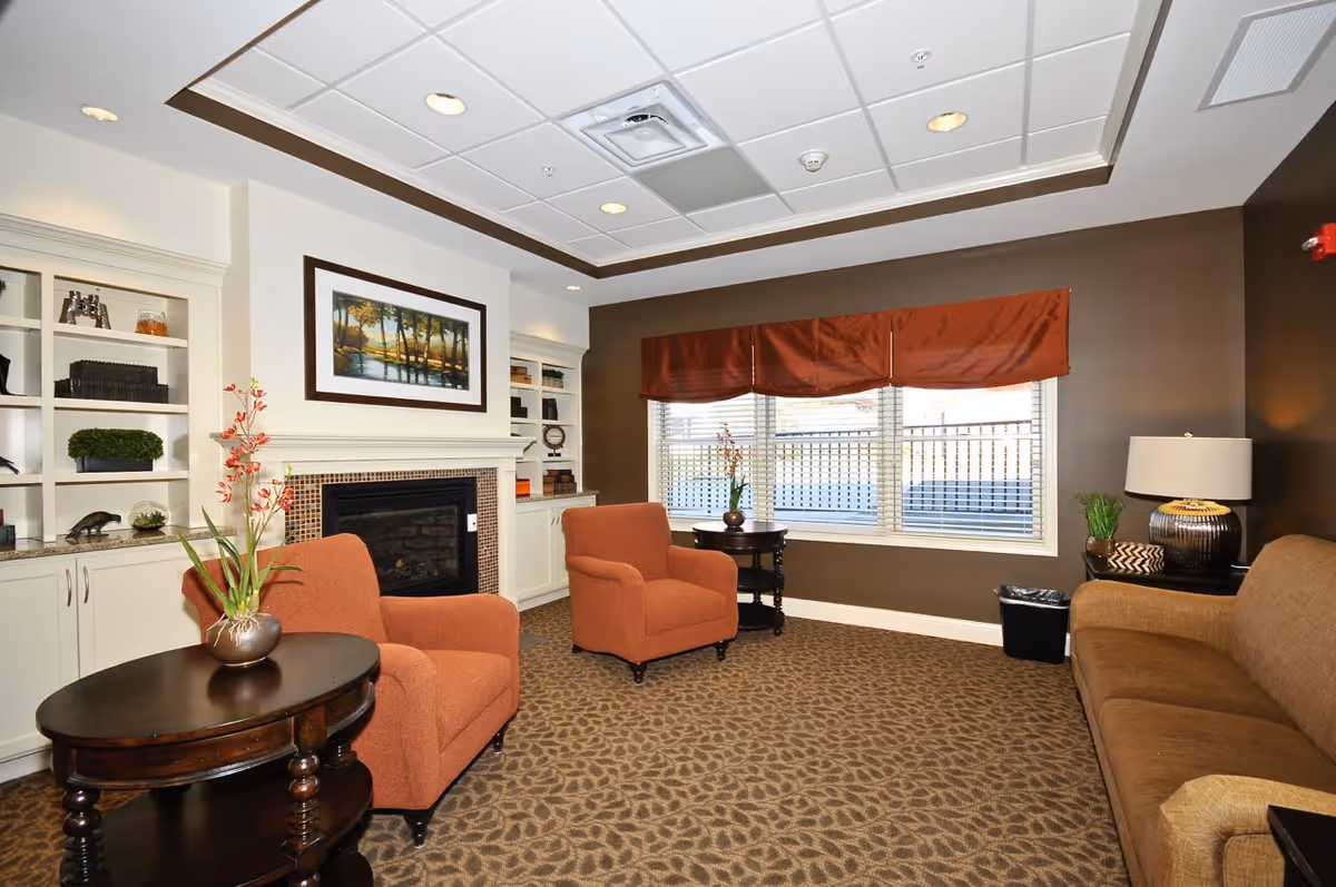 A cozy senior living room with two orange armchairs, a brown sofa, a dark wooden round table with a flower vase, a fireplace with a framed landscape painting above it, built-in white shelves with decorative items, a window with brown valance curtains, and a table lamp on a side table.