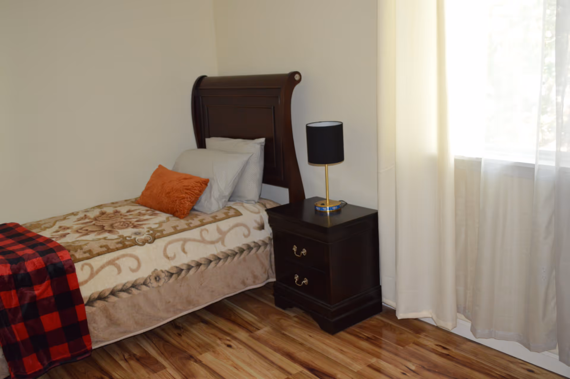 A small bedroom with a single bed featuring a floral patterned blanket, an orange pillow, and a red and black checkered throw. Next to the bed is a dark wooden nightstand with two drawers and a black and gold table lamp. Light-colored curtains cover a window on the right side, and the floor is wooden.