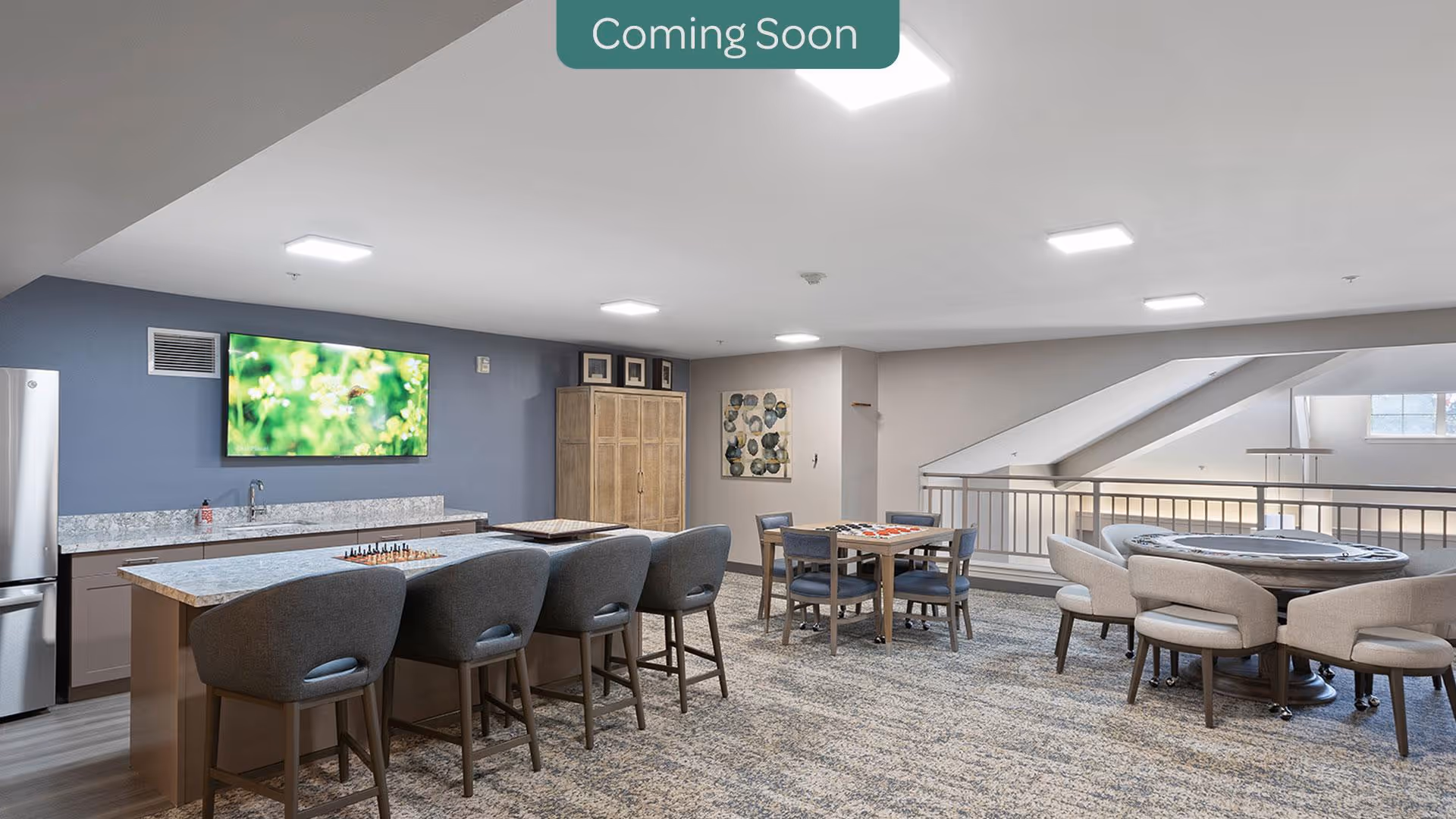 A spacious common area with a kitchen island featuring four bar stools, a wall-mounted TV, a refrigerator, and cabinets. There are two tables with chairs arranged for games or social activities, and a railing overlooking a lower level. The room has modern lighting and neutral decor.