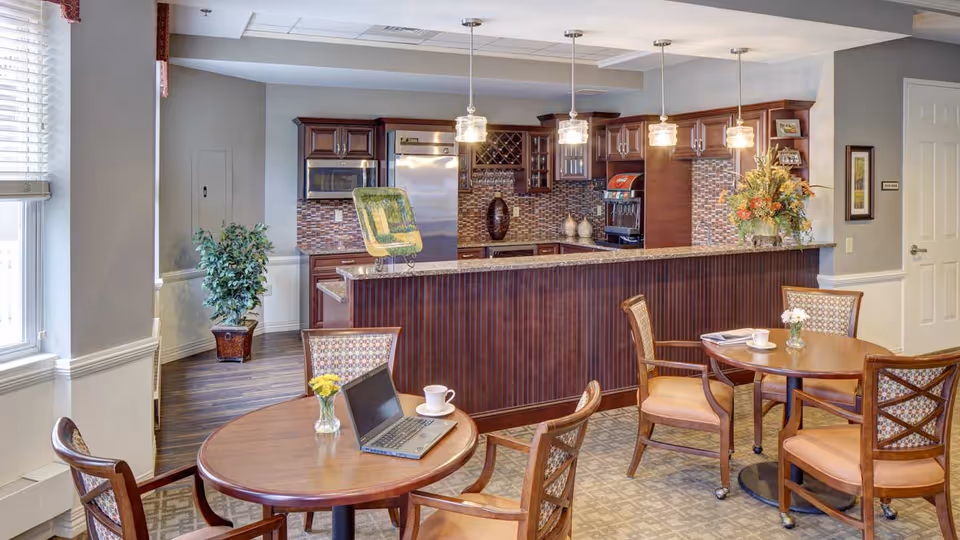 A well-lit communal dining area with round wooden tables and cushioned chairs. One table has a laptop, a cup, and a small vase with yellow flowers. In the background, there is a kitchen area with dark wood cabinets, a stainless steel refrigerator, a microwave, a coffee machine, and pendant lights hanging above a granite countertop. A potted plant is placed near a window with blinds.