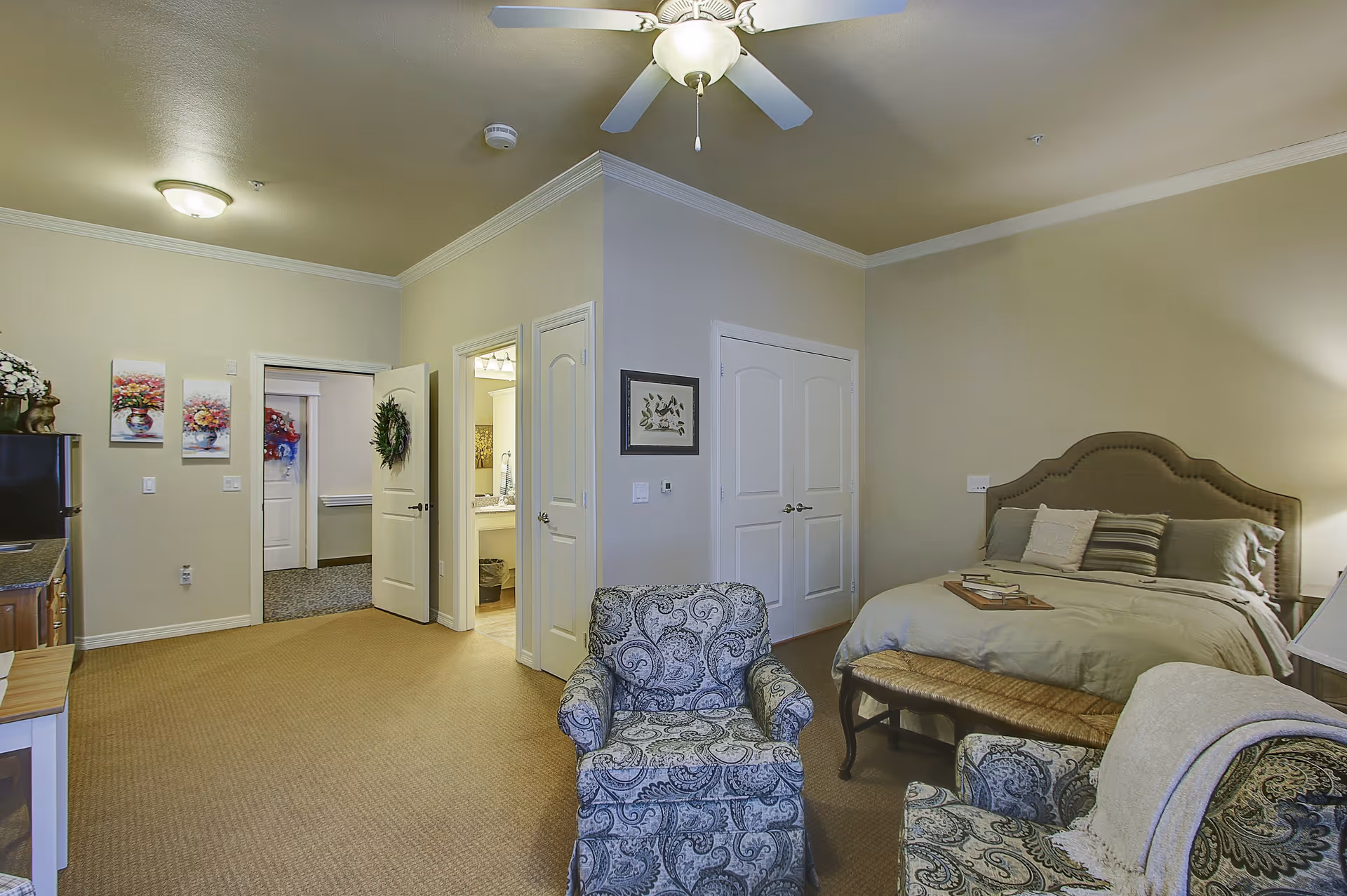 A cozy senior living bedroom featuring a large bed with multiple pillows, two patterned armchairs, a ceiling fan with light, and a small kitchenette area with a mini fridge and cabinets. The room has beige walls and carpet, with two doors leading to other areas, one of which opens to a bathroom with a sink and mirror visible.