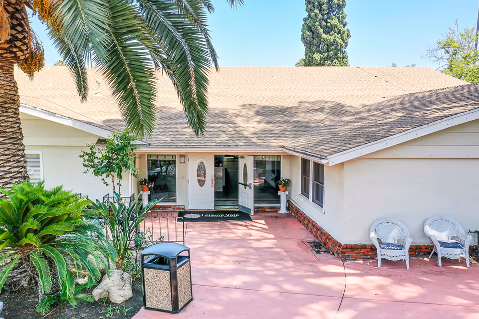 Front exterior view of a single-story building with a light-colored roof and walls, featuring a double-door entrance with glass panels. There are two white wicker chairs with cushions on the right side of the entrance, various green plants including a palm tree on the left, and a trash bin near the walkway. The sky is clear and blue.