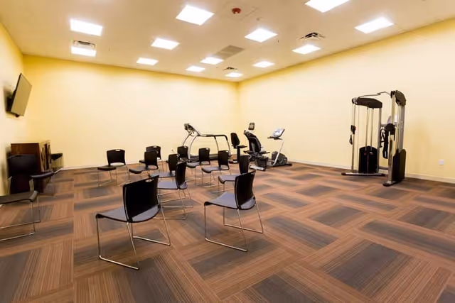 A spacious room with beige walls and a patterned brown carpet floor, containing several black chairs arranged in a semi-circle, two exercise machines including a treadmill and a recumbent bike, and a multi-function weight machine in the corner. A wall-mounted TV is visible on the left side.