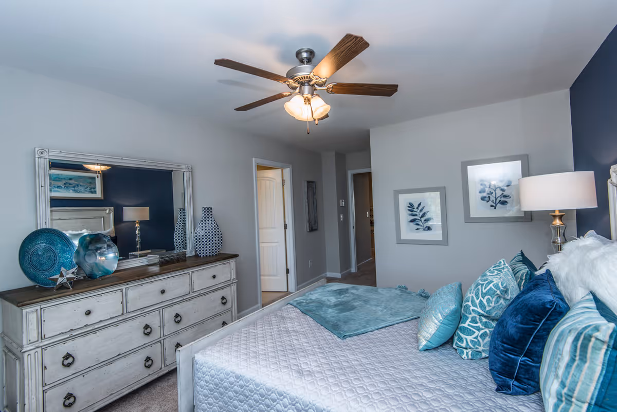 A bedroom with a bed covered in a white quilt and multiple blue and teal pillows. A teal throw blanket is folded at the foot of the bed. On the right side, there is a bedside table with a lamp. On the left side, there is a white dresser with a large mirror above it, decorated with blue and teal vases and decorative plates. The walls are painted light gray and dark blue, with framed botanical artwork hanging. A ceiling fan with lights is mounted on the ceiling.