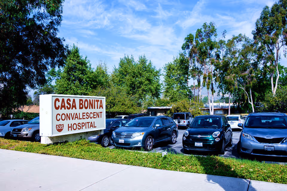 Parking lot with several cars and a large sign reading "Casa Bonita Convalescent Hospital" in front of trees.