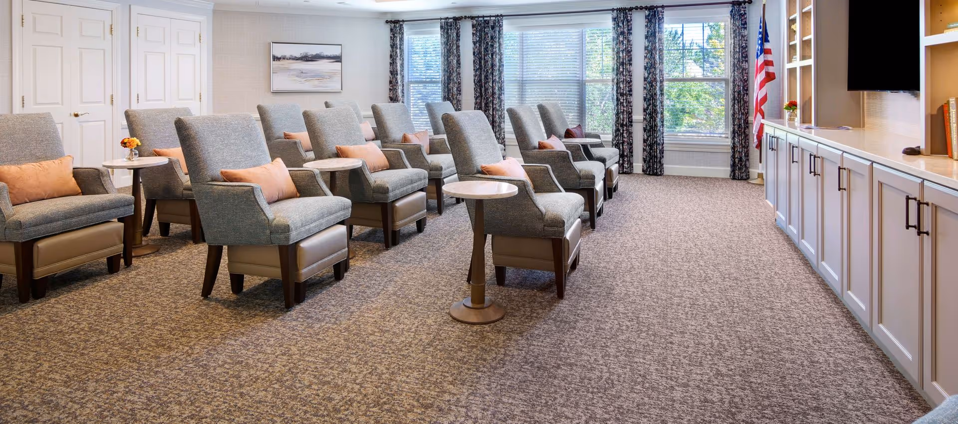 Bright communal lounge with multiple upholstered armchairs and small side tables arranged facing built-in cabinets and large windows.