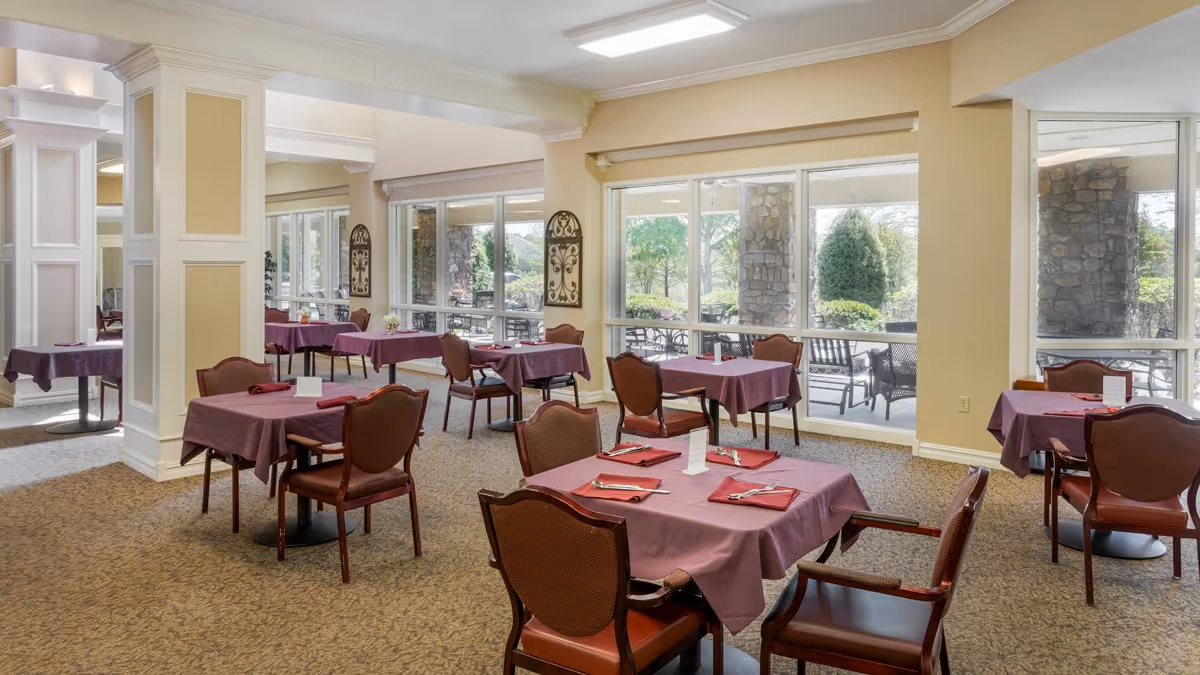 A dining room with several tables covered in purple tablecloths, each set with red napkins and silverware. The room has large windows letting in natural light and offering a view of an outdoor patio area with stone pillars and greenery. The walls are painted a soft yellow and the carpet is a neutral tone.