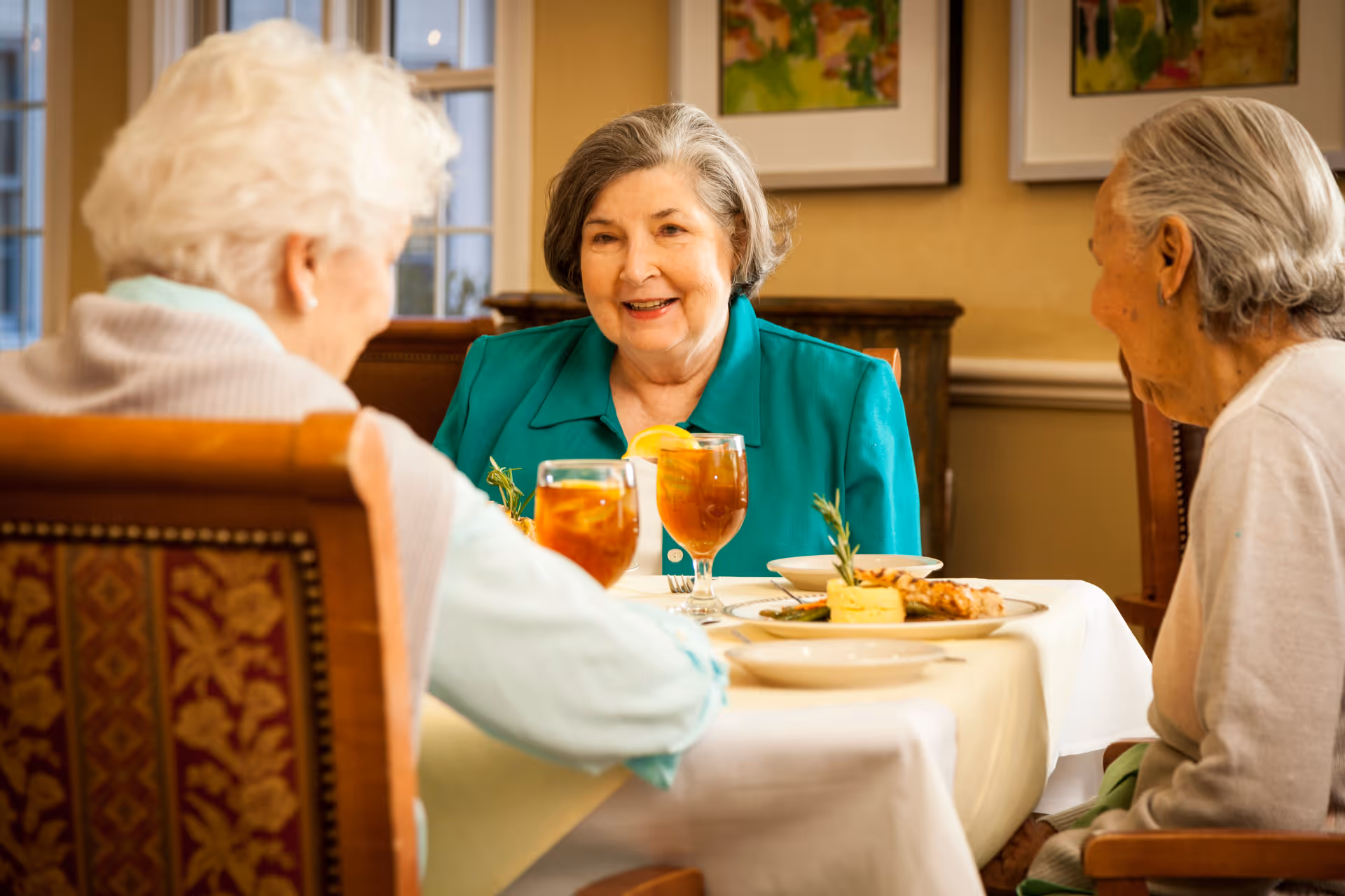 Three elderly women sitting around a dining table in a well-lit room, enjoying a meal and iced tea, engaged in conversation and smiling.