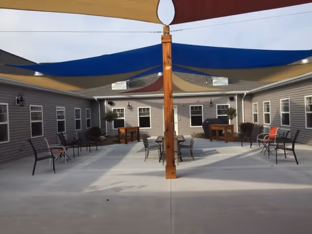 Outdoor courtyard area at Vicinia Gardens Luxury Retirement Living - Intermediate Care, featuring a concrete patio with several chairs and tables arranged under colorful shade sails attached to a central wooden post. The courtyard is surrounded by a single-story building with multiple windows and doors.
