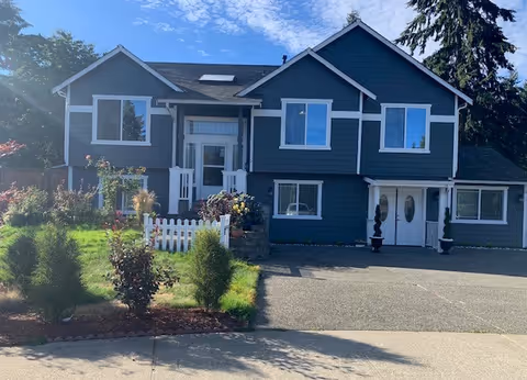 Front exterior view of a two-story dark blue house with white trim, a small white picket fence, landscaped garden with shrubs and flowers, and a paved driveway under a partly cloudy sky.