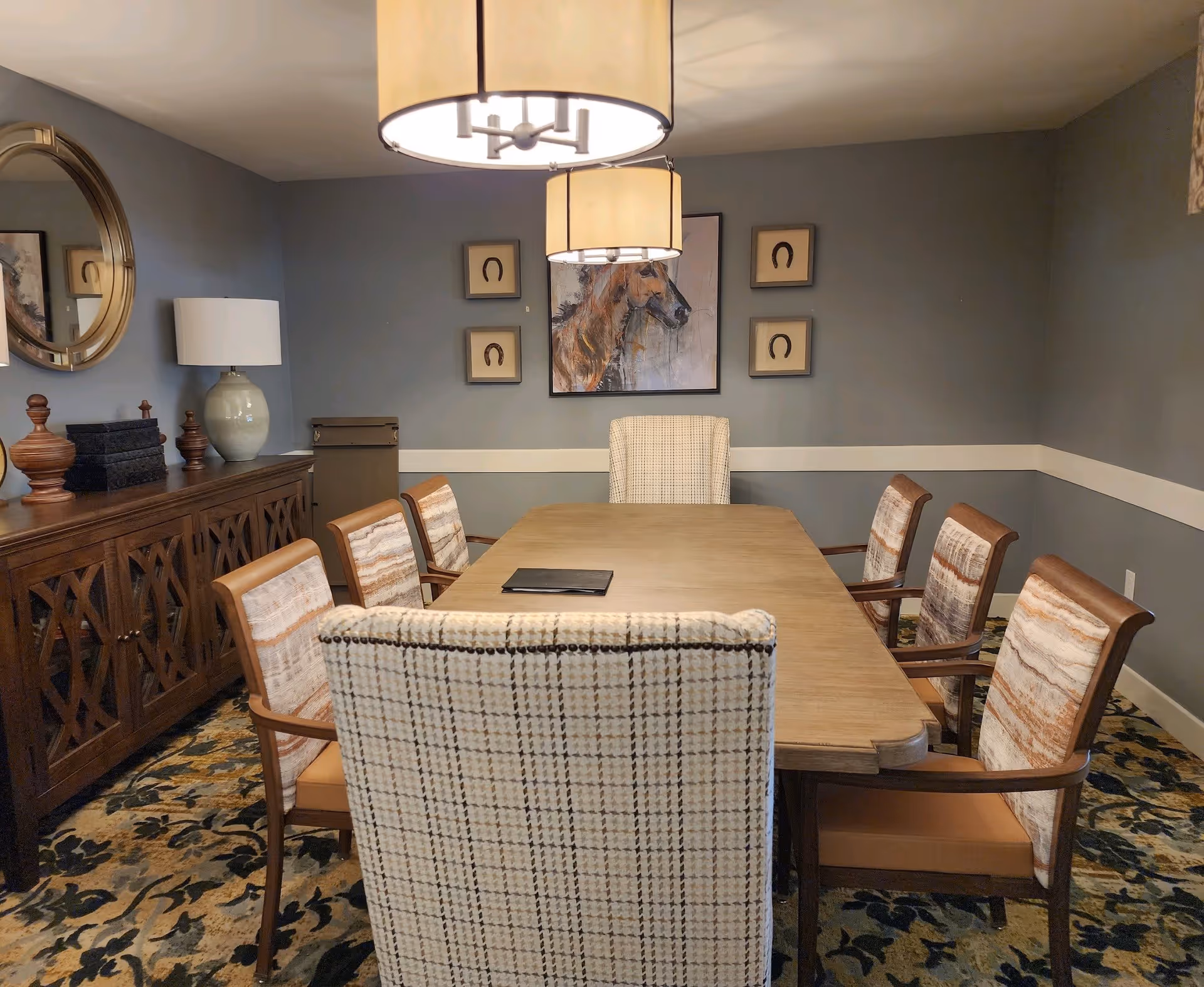 A dining room with a rectangular wooden table surrounded by eight upholstered chairs. The walls are painted gray with a white chair rail. On the far wall, there is a large painting of a horse flanked by four smaller framed pictures of horseshoes. A wooden sideboard with decorative items and a lamp is on the left side of the room. Two large pendant lights hang from the ceiling above the table. The floor is covered with a patterned carpet.