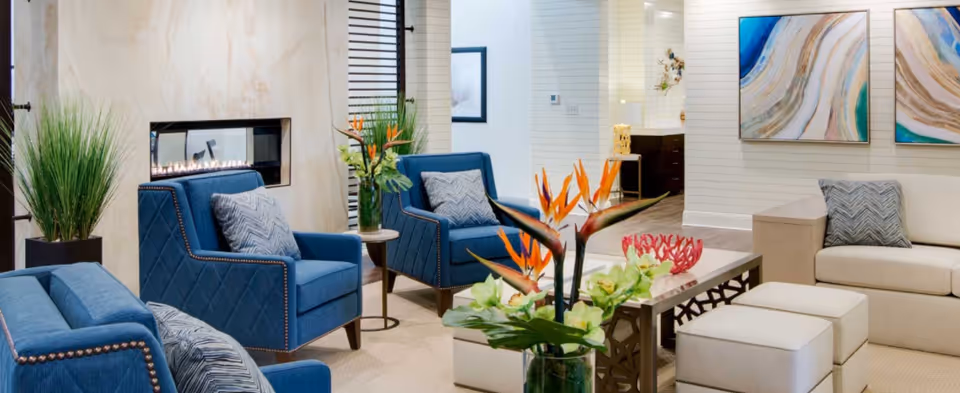 A modern living room with blue upholstered armchairs and a beige sofa with matching ottomans. There is a rectangular coffee table with floral arrangements, including bird of paradise flowers. A built-in fireplace is set in a light-colored wall, and two abstract paintings hang on the wall above the sofa. The room has a bright and clean design with decorative plants and pillows.