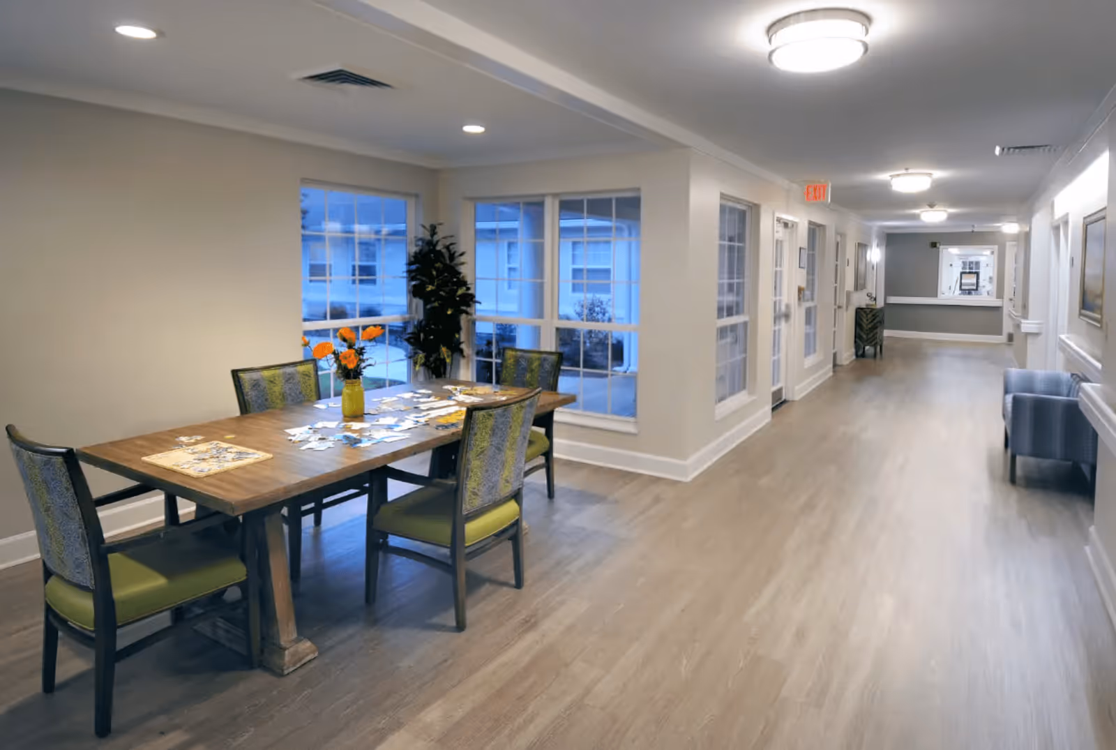 A well-lit hallway in a senior living facility with light wood flooring and beige walls. On the left side, there is a wooden table with four chairs, a vase with orange flowers, and some scattered papers or cards on the table. Large windows behind the table show an exterior view. The hallway extends to the right with additional seating and decor, including a blue armchair and framed pictures on the walls.