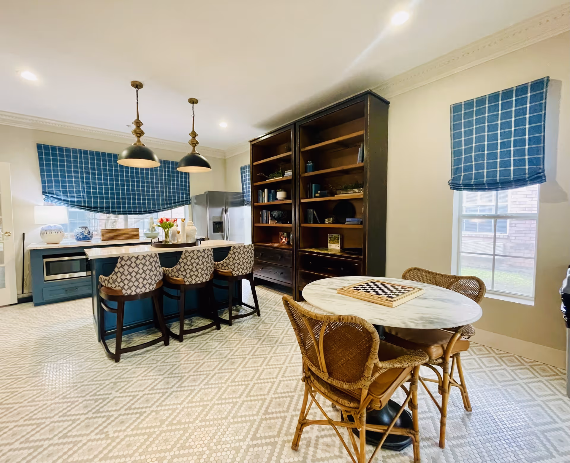 A bright kitchen area with a patterned tile floor, a kitchen island with three patterned chairs, two pendant lights hanging above the island, a stainless steel refrigerator, and a large dark wooden bookshelf. There is also a round marble table with a chessboard on it and two wicker chairs. Blue checkered window shades cover the windows.