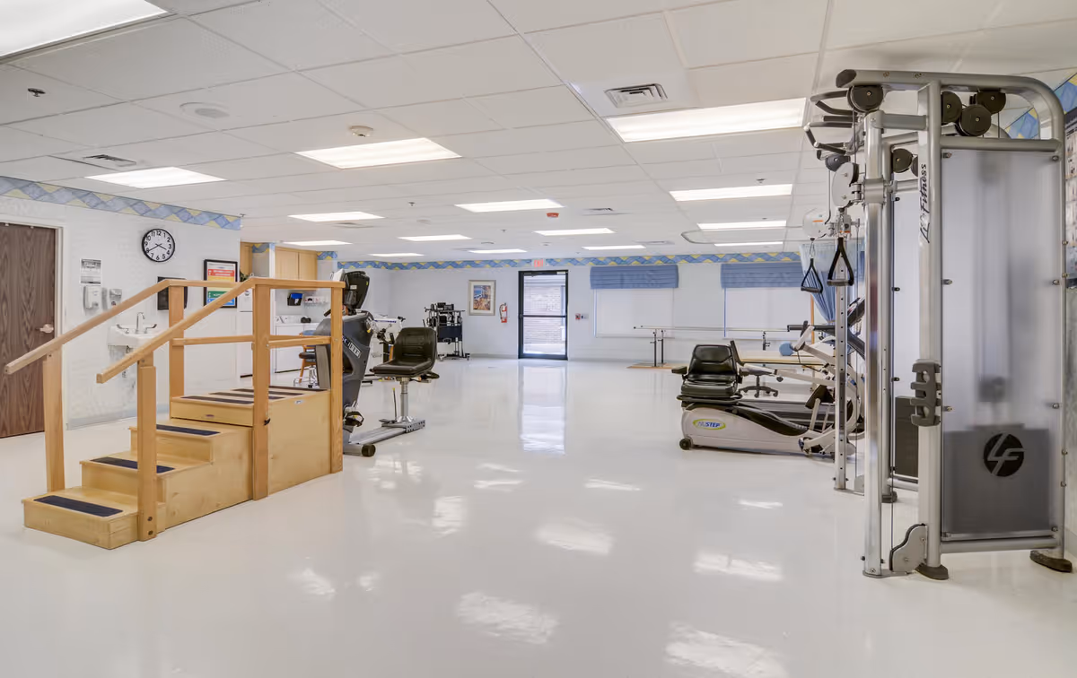 A spacious physical therapy or exercise room with various fitness equipment including a multi-functional exercise machine, stationary bikes, and a wooden staircase with handrails for rehabilitation exercises. The room has a white floor, white ceiling with fluorescent lights, and light-colored walls with a blue and yellow border near the ceiling. There is a clock on the wall and a door at the far end of the room.