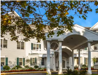 Exterior view of a multi-story assisted living facility building with white siding, green shutters, and a covered entrance supported by columns. Trees with green leaves are visible in the foreground under a clear blue sky.