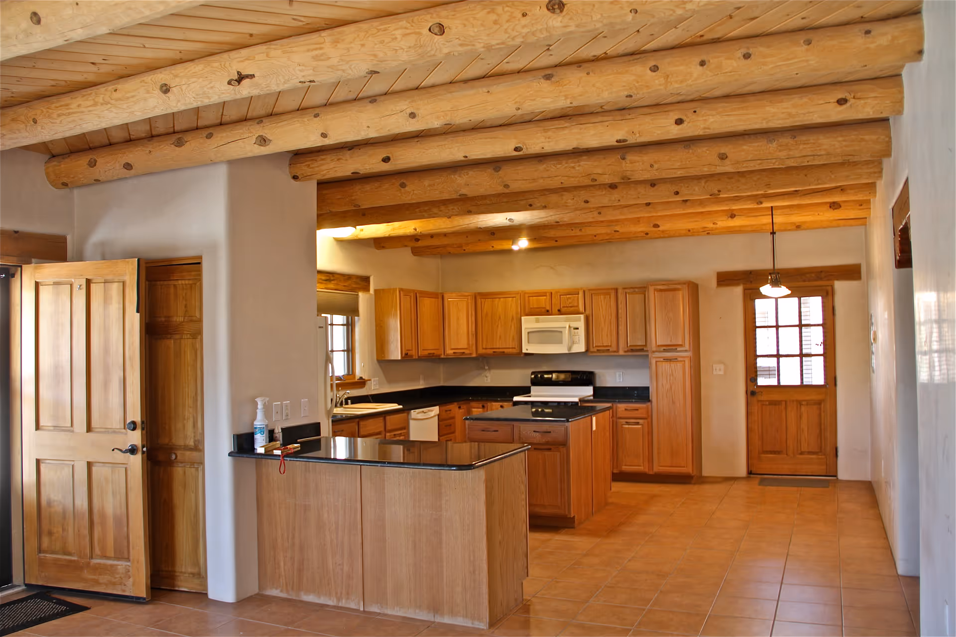 Interior view of a kitchen with wooden cabinets, a black countertop island, a white microwave above a stove, a refrigerator, and a wooden door. The ceiling features exposed wooden beams and the floor is tiled.