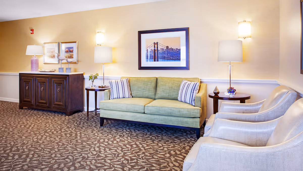 A cozy living room area with a light green sofa adorned with two striped pillows, two beige armchairs, two round wooden side tables with lamps and decorative items, a dark wooden cabinet with a pink lamp and framed pictures above it, and a framed picture of the Golden Gate Bridge on the wall.