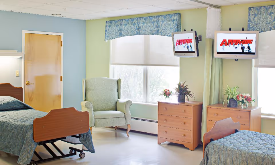 A bright and clean bedroom in a senior living facility featuring two hospital-style beds with blue bedspreads, a green upholstered armchair near a large window with blue patterned valances, two wooden dressers with plants and flowers on top, and two wall-mounted televisions displaying the word 'AMAZE'.