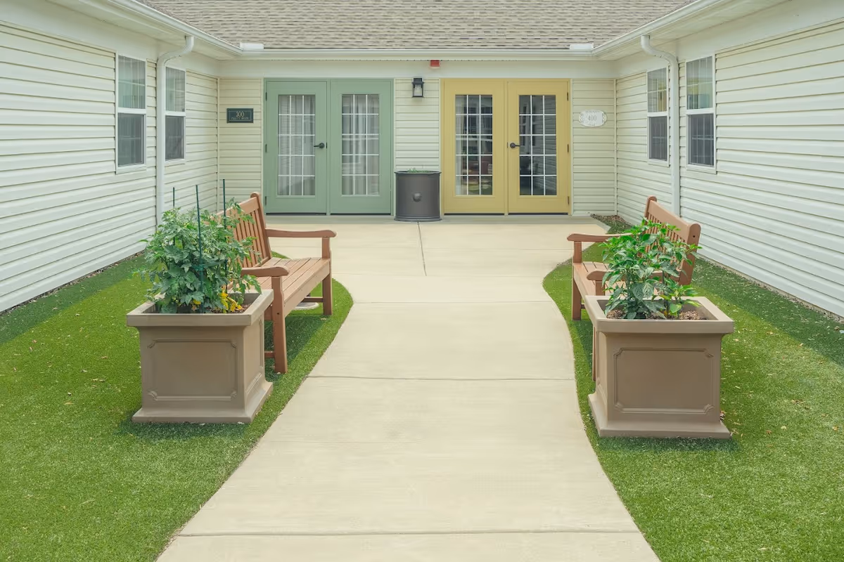 Outdoor courtyard area with two wooden benches facing each other, each accompanied by a large planter with green plants. The courtyard is surrounded by light-colored siding walls with multiple windows and two sets of French doors painted in green and yellow. The ground is a combination of concrete walkway and artificial grass.