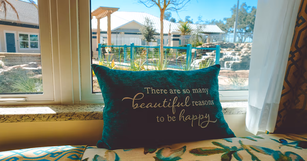 A decorative pillow with the text 'There are so many beautiful reasons to be happy' placed on a cushioned window seat. Outside the window, there is a view of a garden area with a waterfall feature, plants, and a building in the background under a clear blue sky.
