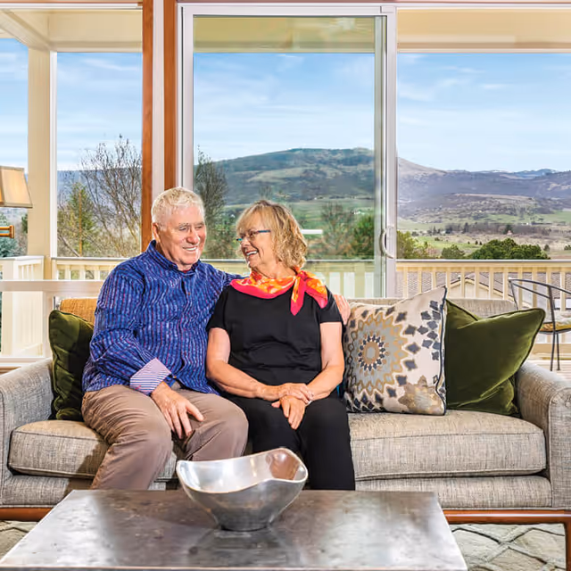 An elderly couple sitting closely together on a beige sofa in a bright living room with large windows showing a scenic mountain view. The man is wearing a blue striped shirt and beige pants, and the woman is wearing a black outfit with a colorful scarf. There are decorative pillows on the sofa and a metallic bowl on the coffee table in front of them.