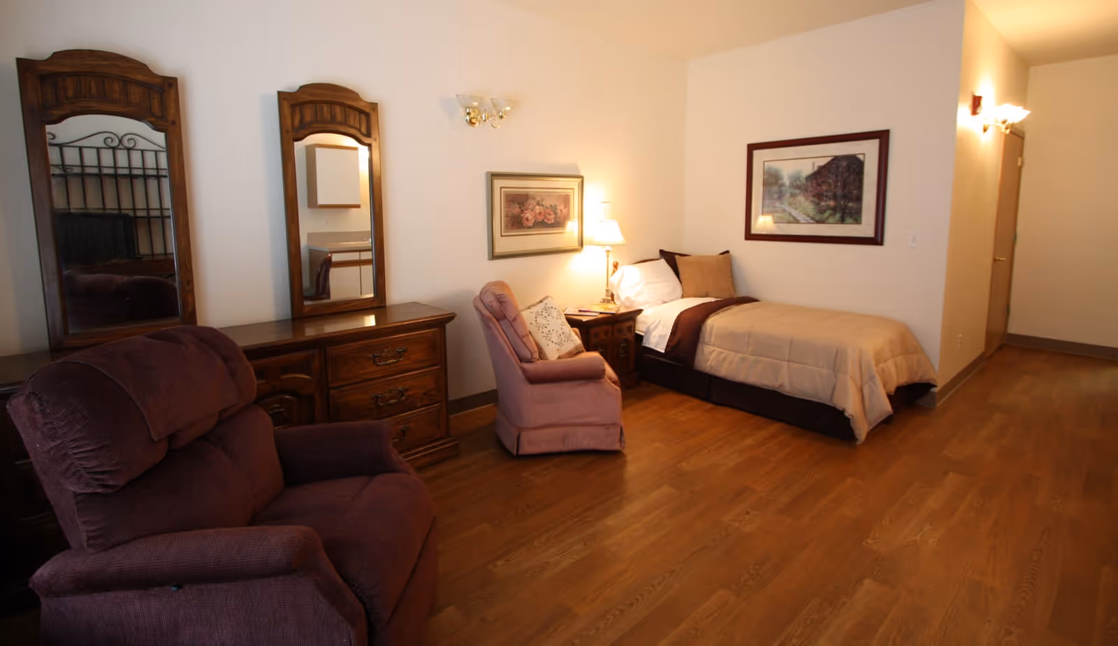 A cozy bedroom in a senior living facility featuring a single bed with beige bedding, a wooden nightstand with a lamp, two framed pictures on the walls, a pink upholstered armchair, a dark purple recliner, and a wooden dresser with two tall mirrors. The room has wooden flooring and soft lighting.