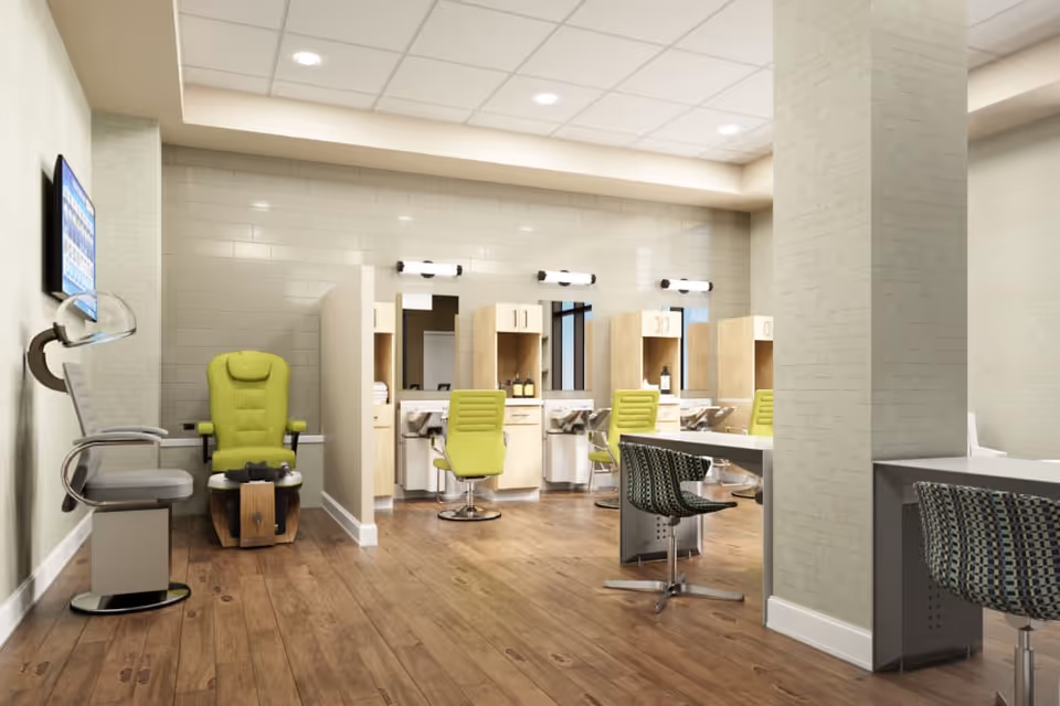 A modern salon area in a senior living facility with light wood flooring, beige tiled walls, and a drop ceiling with recessed lighting. The room features several lime green salon chairs in front of individual stations with mirrors and storage cabinets. There is also a pedicure chair and a couple of patterned swivel chairs near a table on the right side.