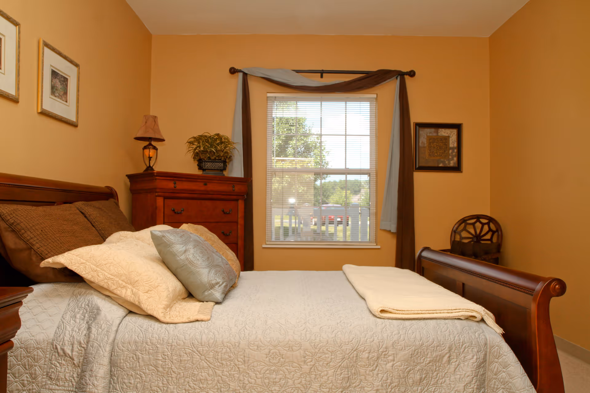 A cozy bedroom with a wooden bed frame, neatly made with a white quilt and multiple pillows. There is a wooden dresser with a lamp and a potted plant on top, a window with blinds and curtains, and framed artwork on the walls. A wooden chair is placed in the corner of the room.